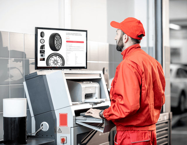 A technician in a tire shop uniform looking at a desktop screen showing a tire product page with size, thumbnails and an add-to-cart button.