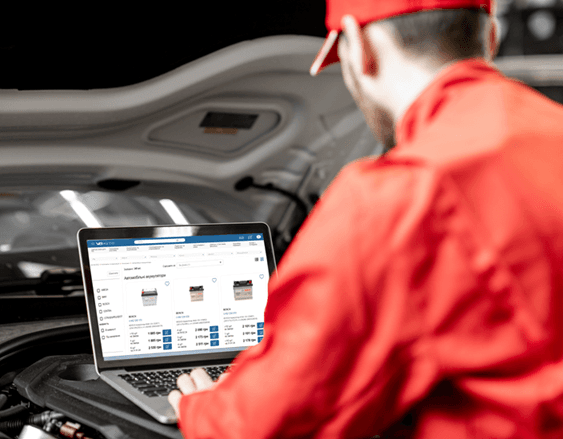 A mechanic in red overalls standing over an open car hood, checking a laptop showing the VBavto parts catalogue with product listings and prices.
