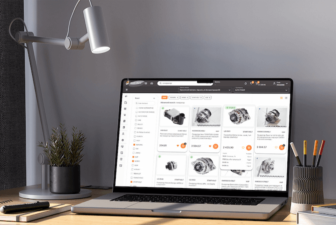 A laptop on a desk showing the Zenit Auto parts catalogue: alternators in a grid with prices and stock marks, brand filters down the left side and a search bar across the top.